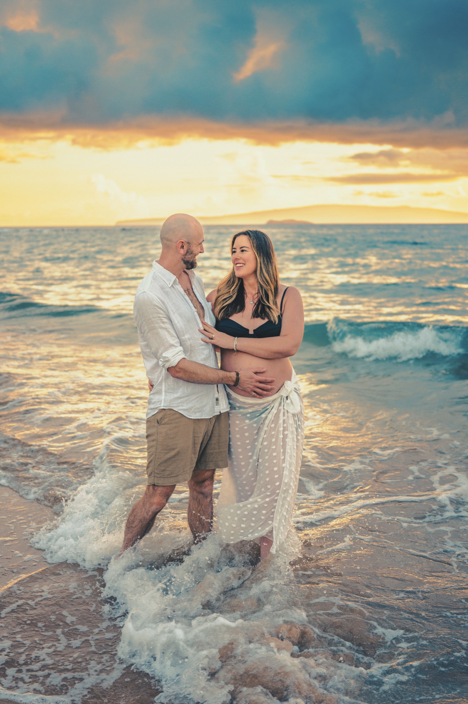 Expectant parents standing together in the ocean at a golden Maui sunset